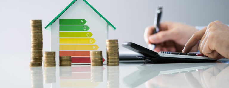 Mit einem Taschenrechner wird eine Ersparnis ausgerechnet und notiert. Es türmen sich Münzen vor einem Haus mit schematischer Darstellung von Energieeffizienzklassen.